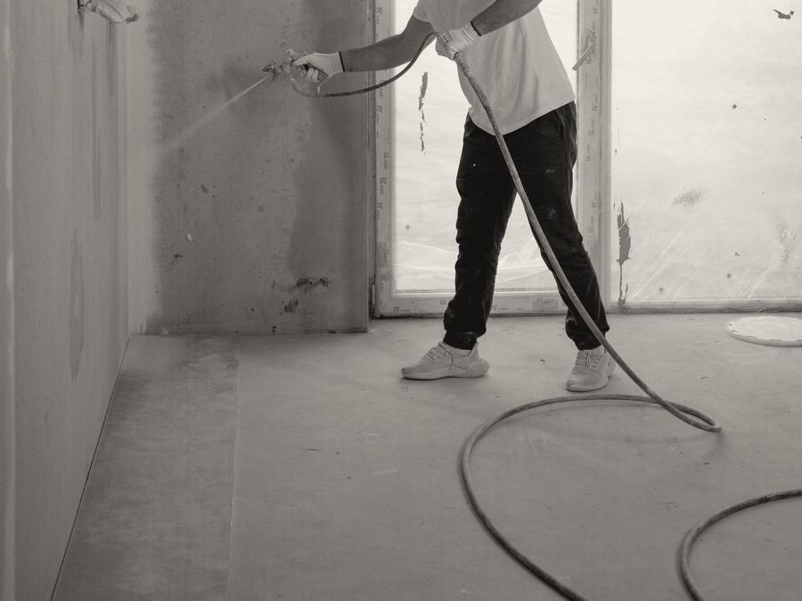 Sample photo: a painter spraying an interior wall with an airless sprayer, hose coiled on the floor